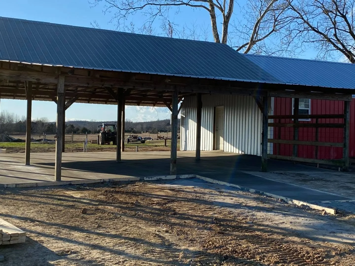 A partially constructed building on a farm with a metal roof, wooden support beams, and a red and white siding wall. In the background, there's a tractor, open fields, and leafless trees under a clear blue sky.