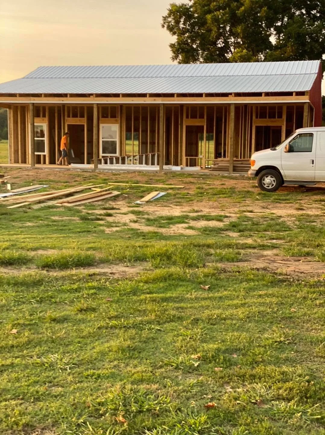 Construction site of a house with a metal roof, wooden framing, and two windows. A worker is walking on the left side of the building. Construction materials are scattered on the ground in front, and a white van is parked to the right. There are gree