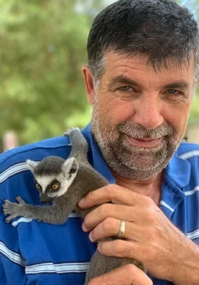 A man with dark hair and a beard holding a small lemur with big eyes and a long tail outdoors.