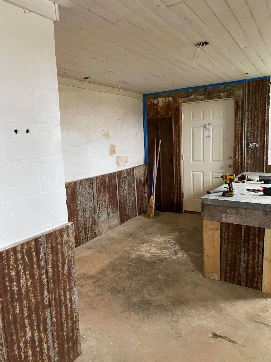 Room under renovation with exposed walls, construction tools on a makeshift counter, a broom and equipment near a door, and wires hanging from the ceiling.