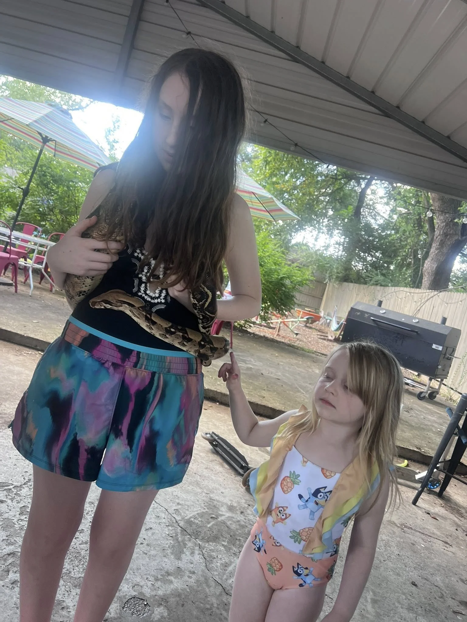 Two young girls standing outdoors under a covered area, one holding a snake, with trees and patio chairs in the background.