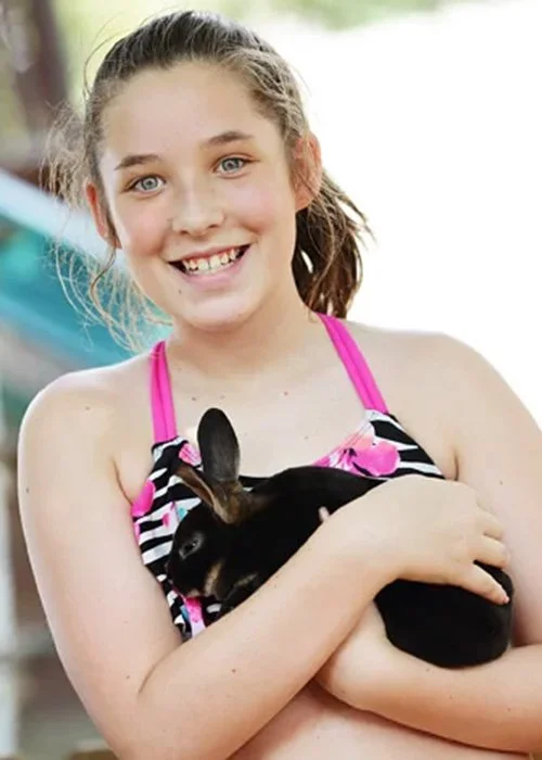 A girl with a ponytail, smiling, holding a black and white rabbit outdoors.