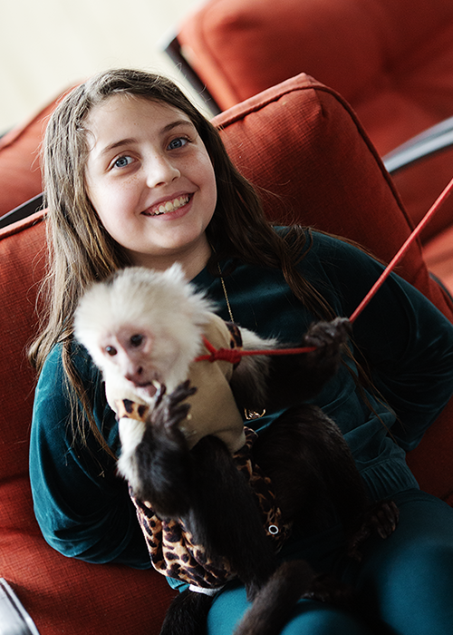 Young girl smiling while holding a small white dog wearing a sweater, sitting on a red sofa.