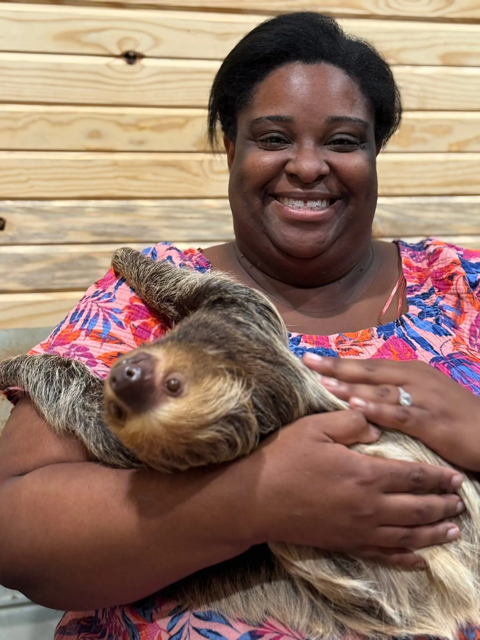 A woman with dark hair smiling and holding a large, cuddly ferret in her arms against a wooden backdrop.