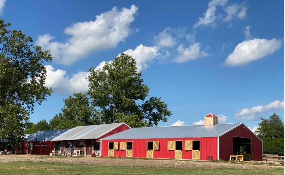 A red barn with a metal roof, multiple windows, and a small tower, situated in a rural area with trees and a blue sky with clouds.
