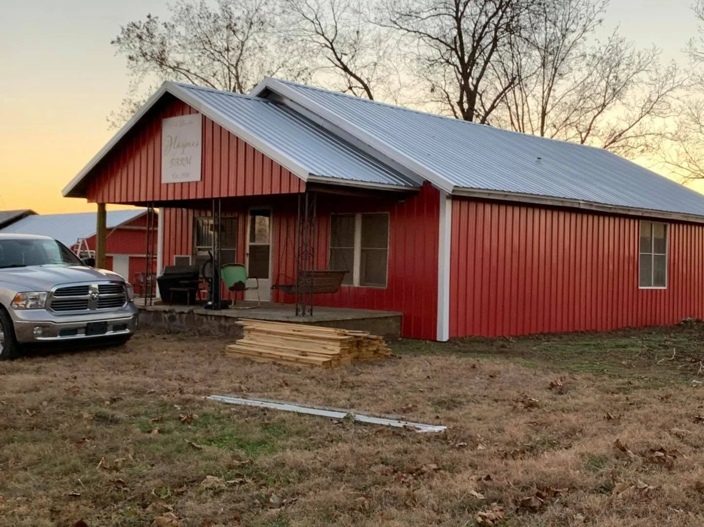 A red house with a metal roof and a front porch, with a stack of wood on the ground nearby, a ladder, and a silver truck parked in the yard during sunset.