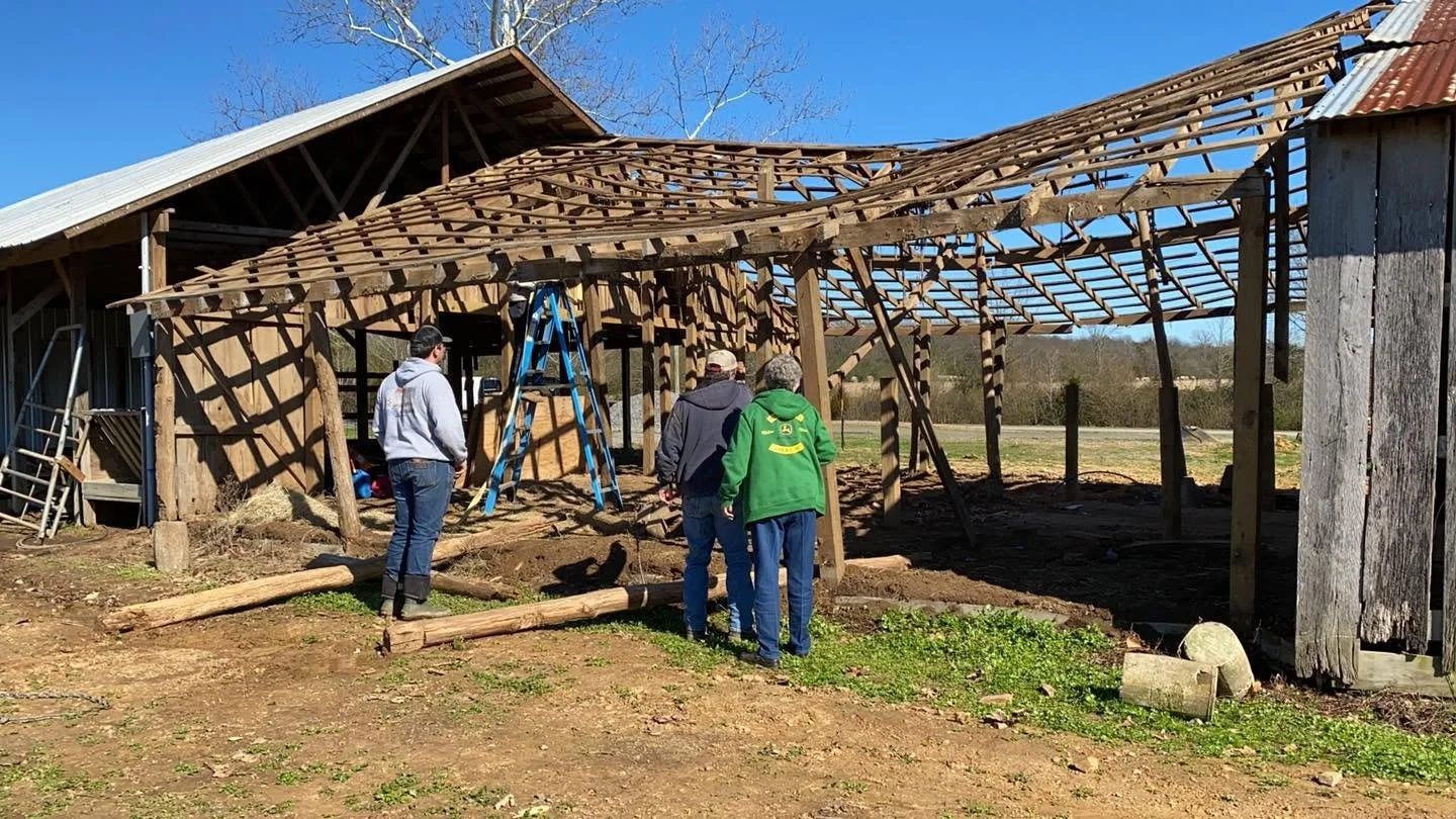 Three people working on the wooden structure of a barn or shed with a damaged roof, one person appears to be on a ladder, in a rural area with open fields and blue sky.