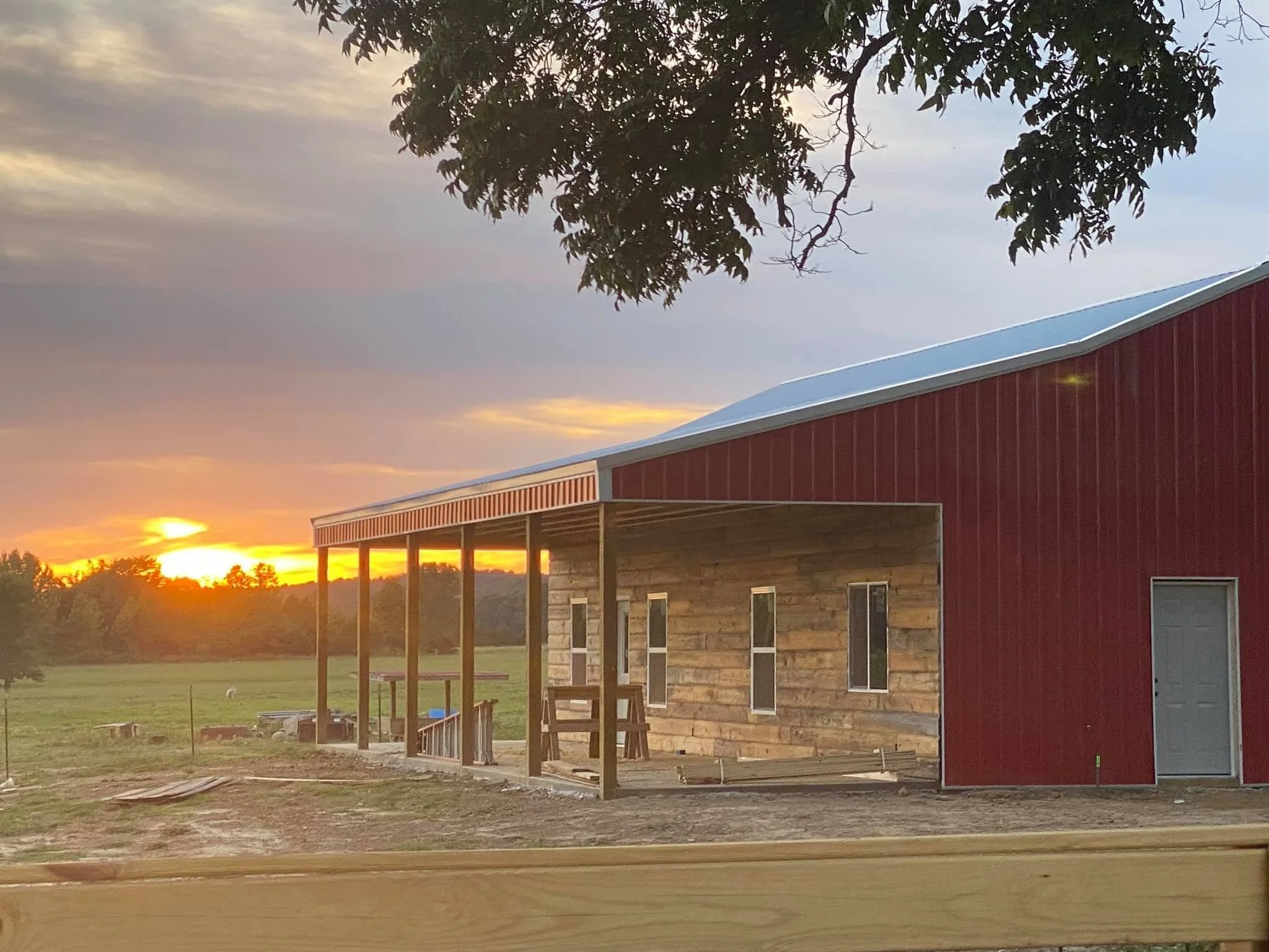A house with a red exterior and a metal roof is under construction, with some wooden structural elements visible. The scene is set during sunset, with a sky filled with clouds and the sun partially visible on the horizon. A large tree with dark leaves is in the foreground.