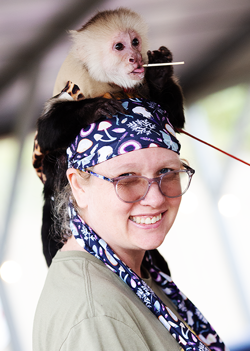 A woman with glasses and a colorful headscarf smiling as a baby capuchin monkey sits on her head, holding a stick in its mouth.