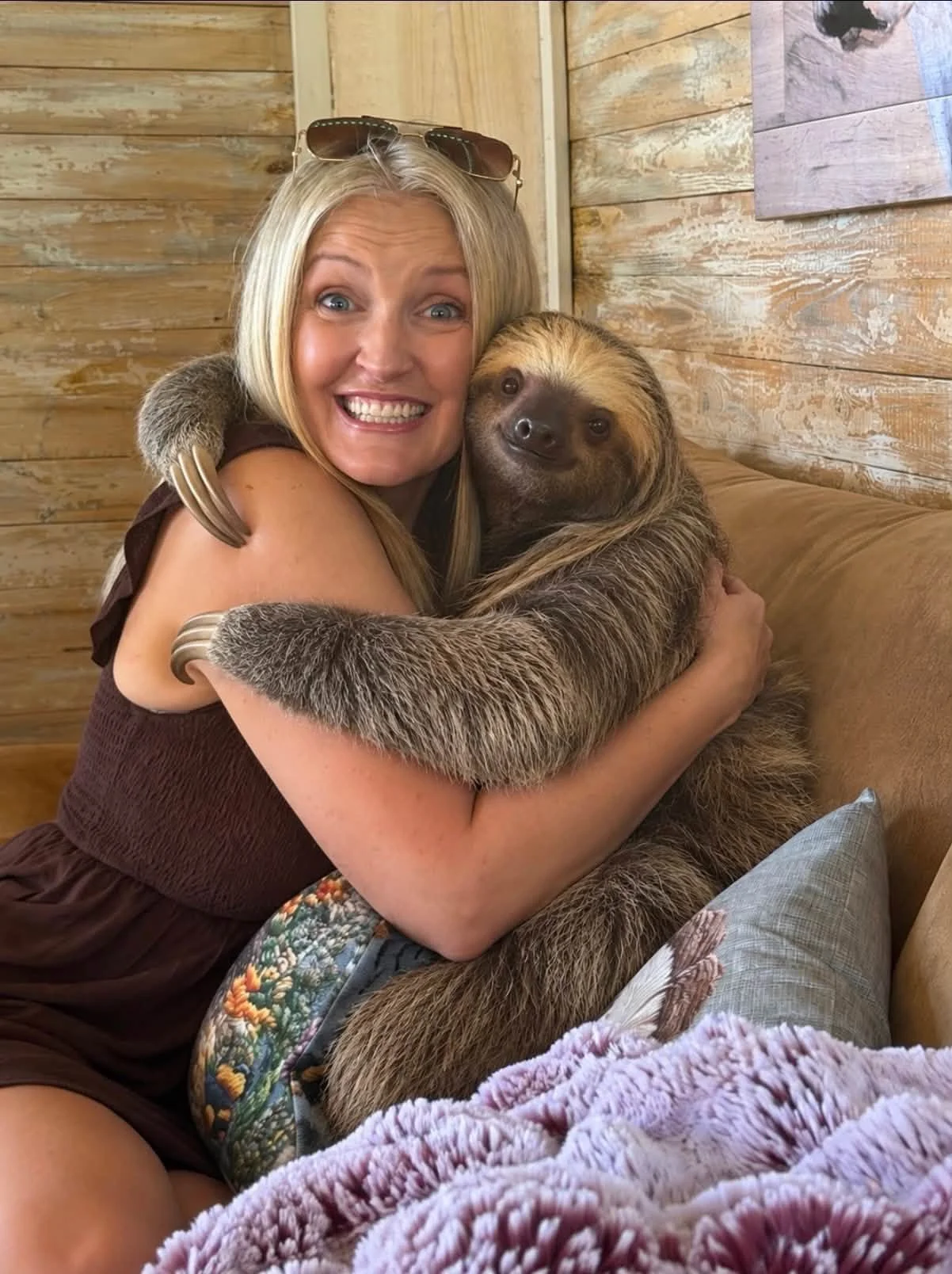 A woman with sunglasses on her head, smiling eagerly, hugging a sloth resting on her lap inside a wooden cabin or room.