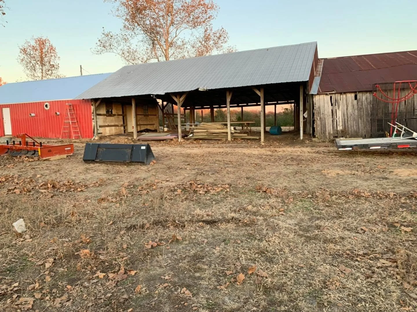 A farm or barn area under construction with wooden and metal structures, construction materials, and equipment scattered on dry ground with leaves.