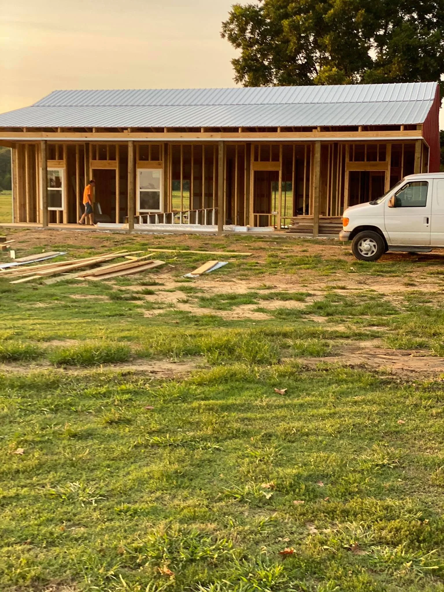 A house under construction with exposed wooden framing, situated on a grassy field with a white van parked nearby, at sunset.