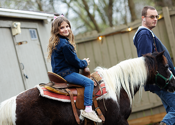 Young girl with long wavy hair riding a brown and white horse, wearing a denim jacket and pink cat ear headband, with a man leading the horse in an outdoor setting.