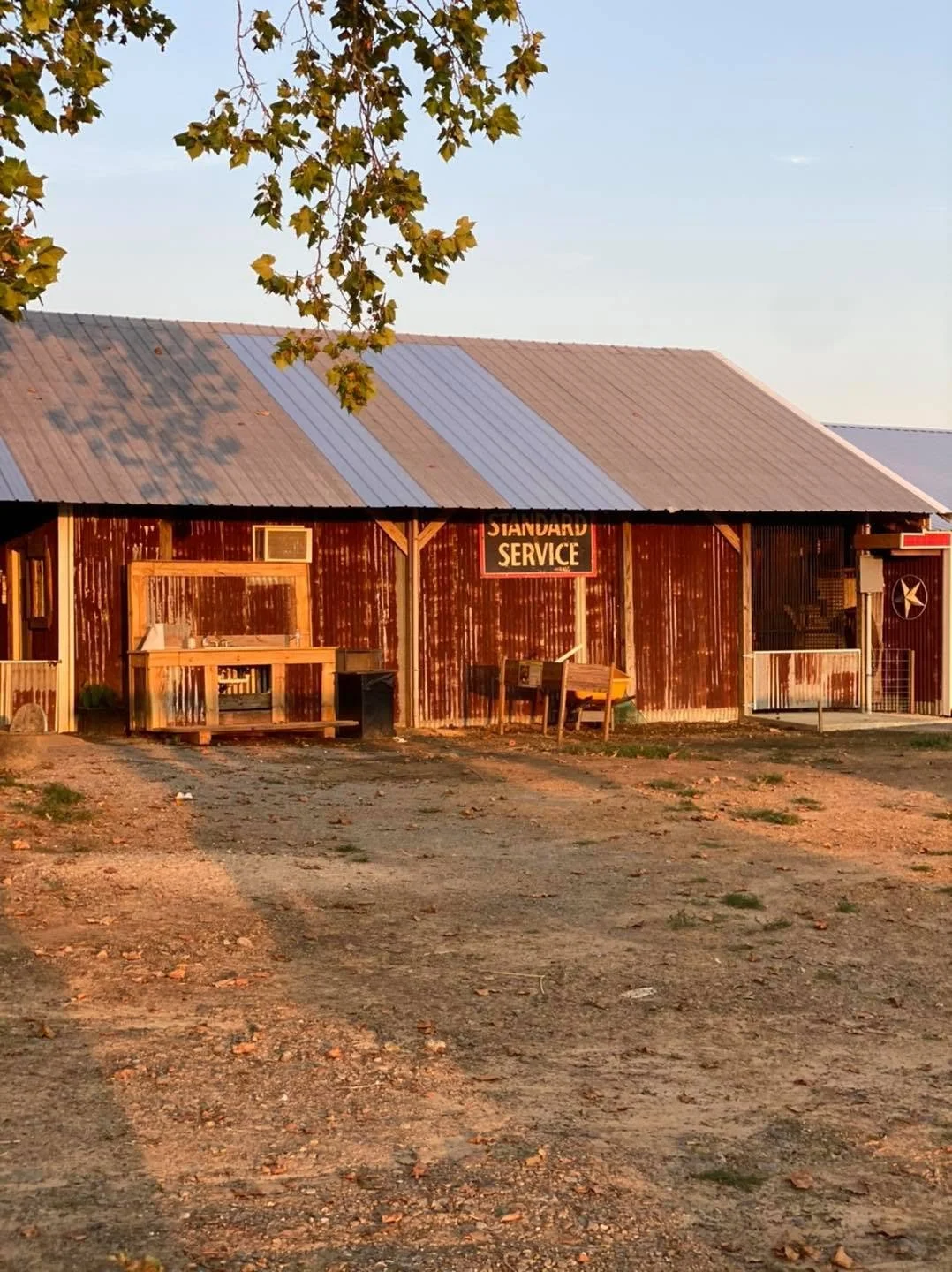 Rustic wooden building with a corrugated metal roof labeled 'Standard Service,' outdoor window, and various objects outside, including a wheelbarrow and a small gate, under a tree with green leaves.