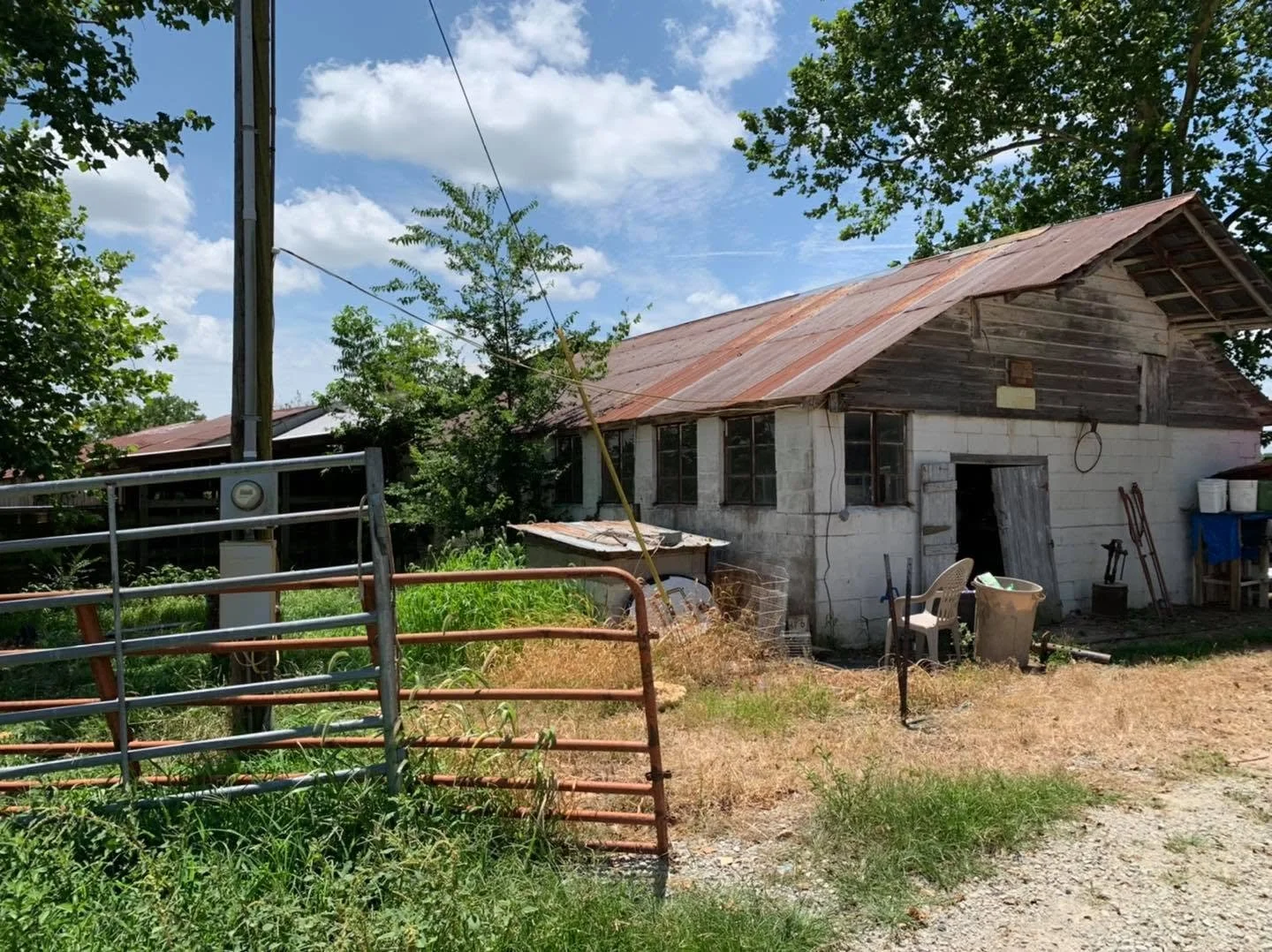 A rustic old building with a rusty metal roof, surrounded by overgrown grass and trees, with various outdoor items scattered around.