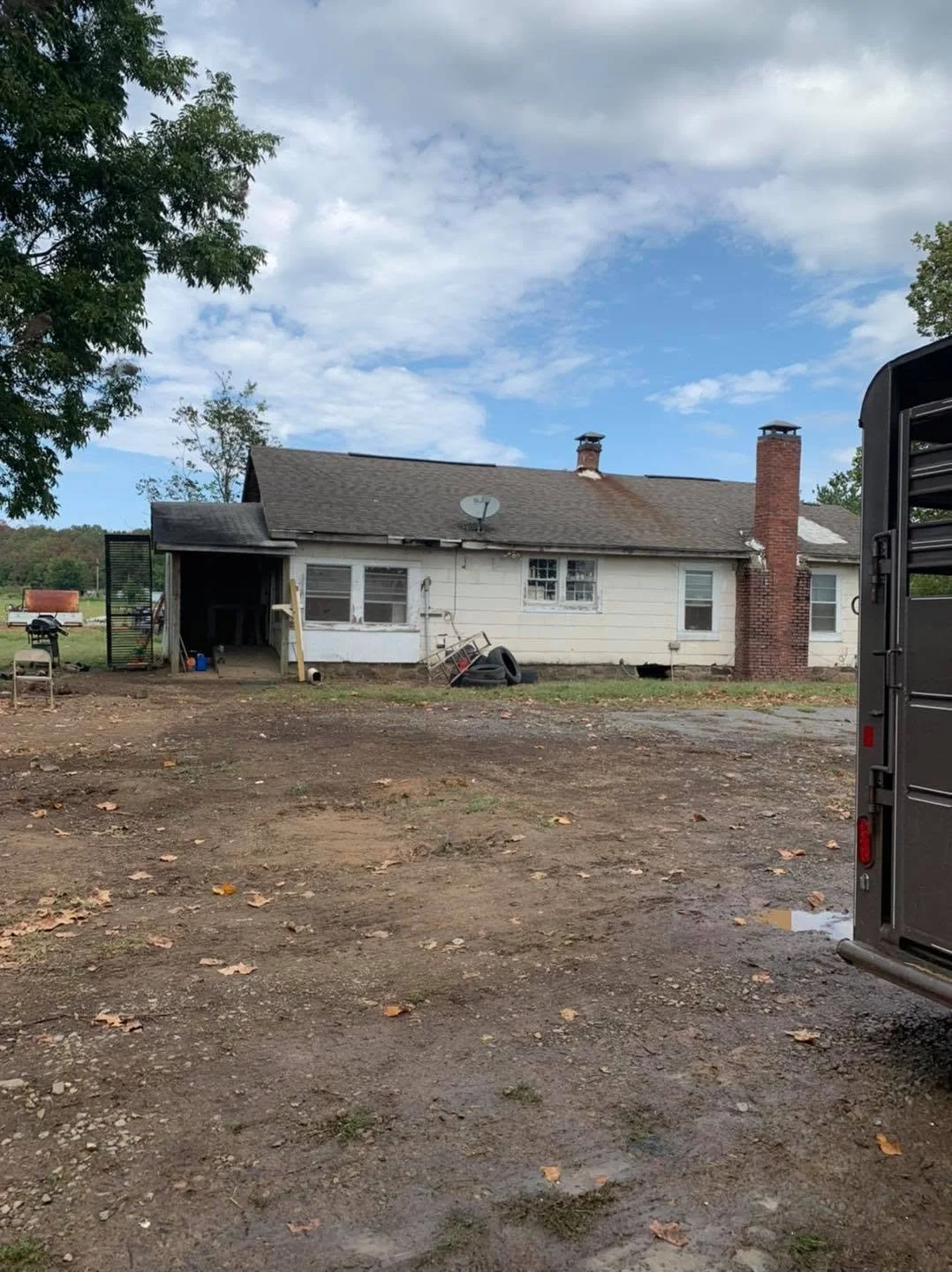 Rear view of a white, single-story house with a brick chimney, satellite dish, and multiple windows. The yard is mostly dirt with leaves, and there are some outdoor items like tires and a chair. Part of a trailer or truck is visible on the right side