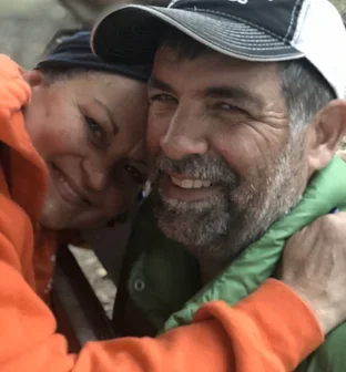 Close-up of a smiling woman and a smiling man hugging each other outdoors.
