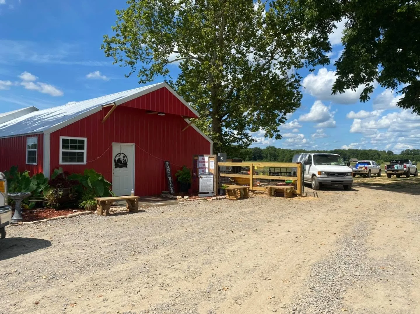 A red barn with white trim under a partly cloudy sky, surrounded by vehicles and outdoor seating, with a gravel area in the foreground.