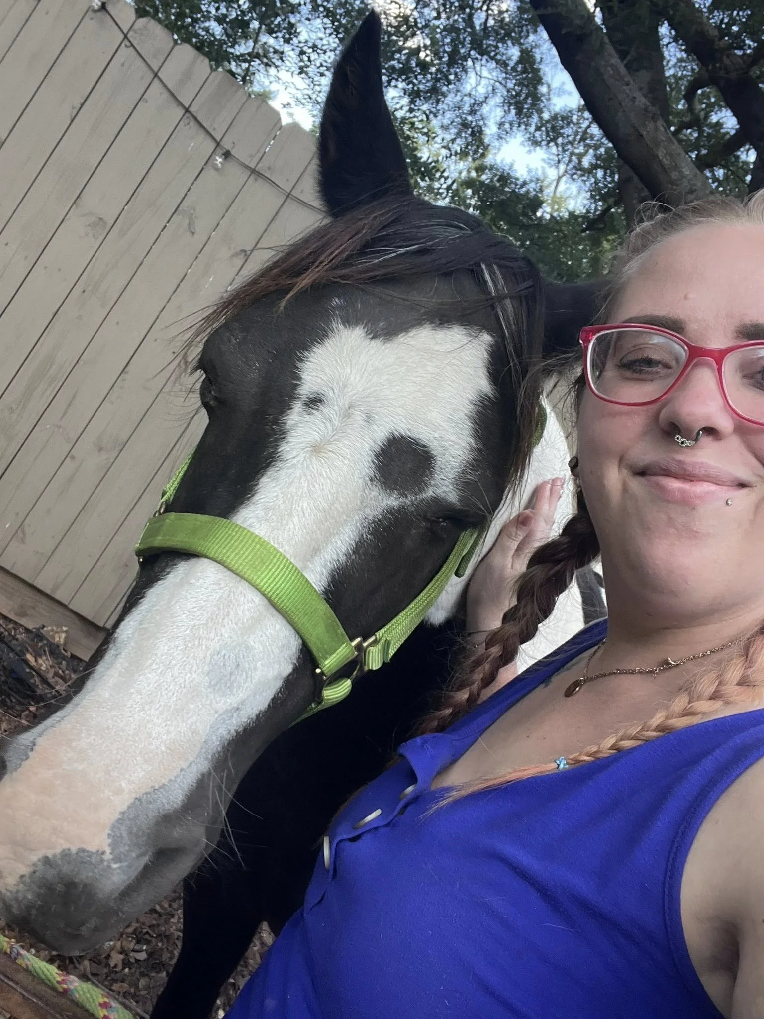 A young woman with braided hair, pink glasses, and a septum piercing taking a selfie with a black and white horse wearing a green halter, in an outdoor setting with trees and a wooden fence.