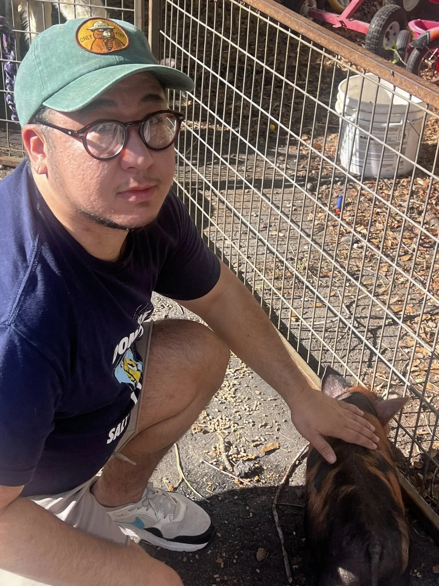 A man wearing glasses, a teal cap with a logo, a dark t-shirt, and light-colored shorts, is crouching down petting a brown and black dog behind a white metal fence. The ground is covered with leaves, and there are some toys and a bucket inside the fe