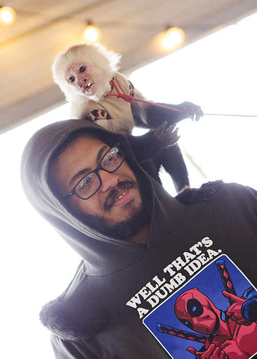 A man with glasses and a hooded sweatshirt smiling as a white monkey wearing a tan jacket sits on his shoulder, holding a leash.