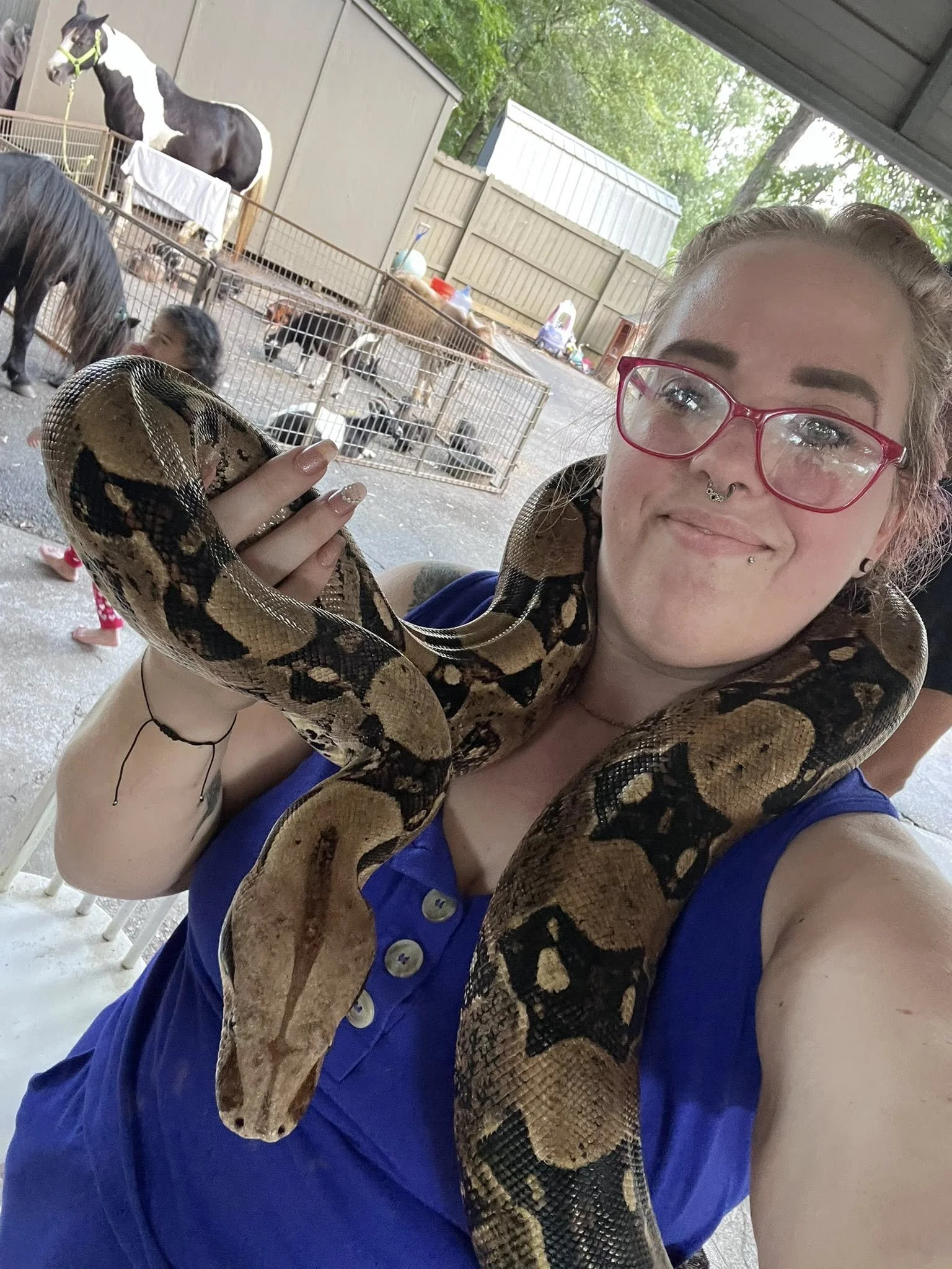 A person with glasses and a piercing on her nose and lip, holding a large snake around her neck, standing in front of a fenced area with small animals and a horse in the background.