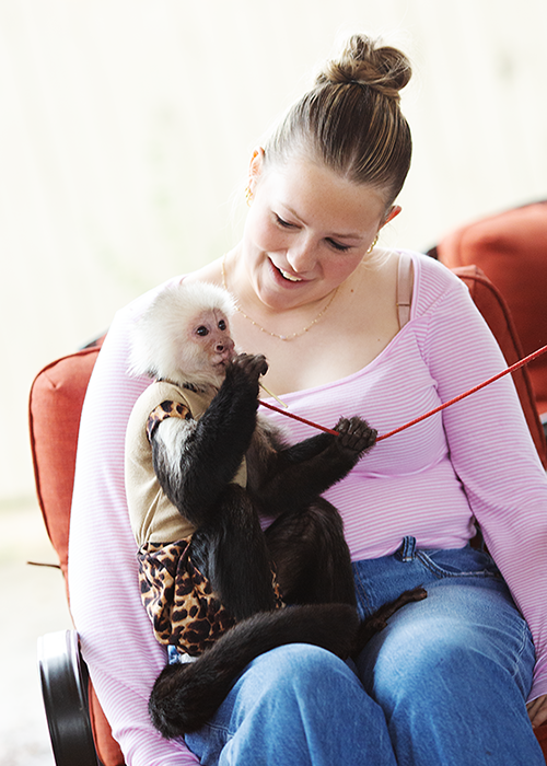 A young woman sitting on a chair holding a small monkey wearing a leopard print outfit, with the monkey holding a red string, in a cozy indoor space.