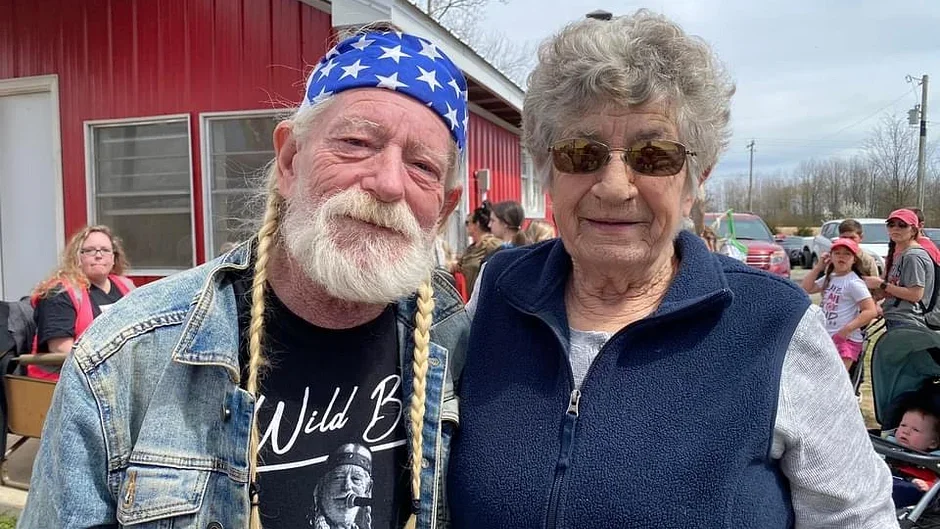 A man with a white beard and braids wearing a bandana with stars, and a woman with curly gray hair and sunglasses, smiling outdoors at an event with children and adults in the background.