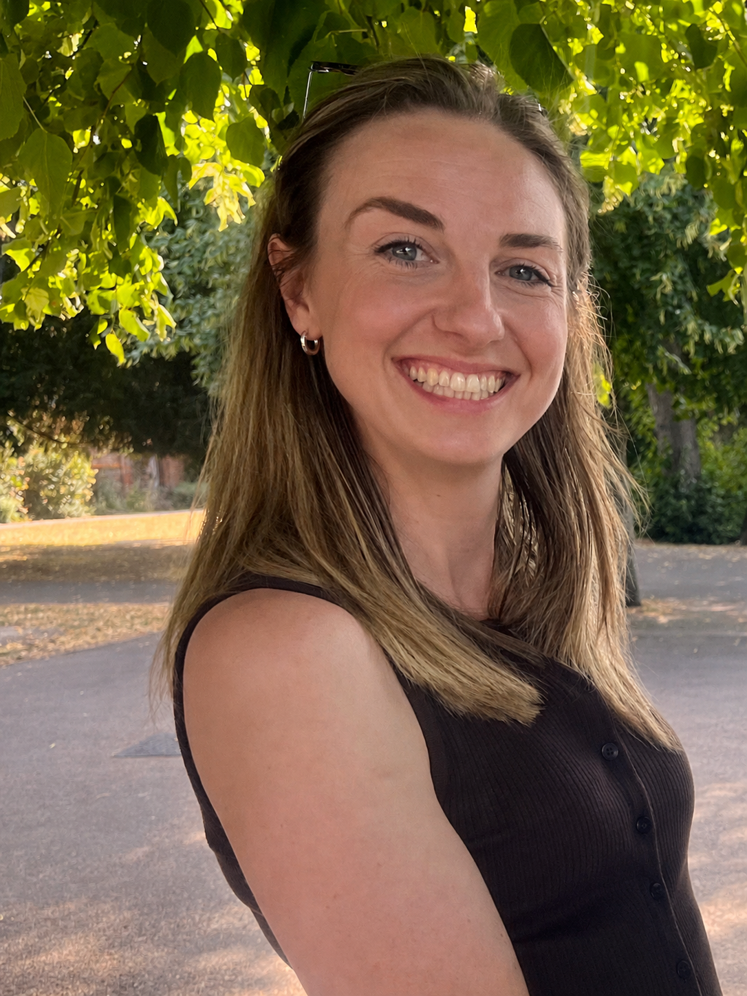 A woman with long, light brown hair, wearing a sleeveless black top, smiling outdoors under a leafy green tree on a sunny day.