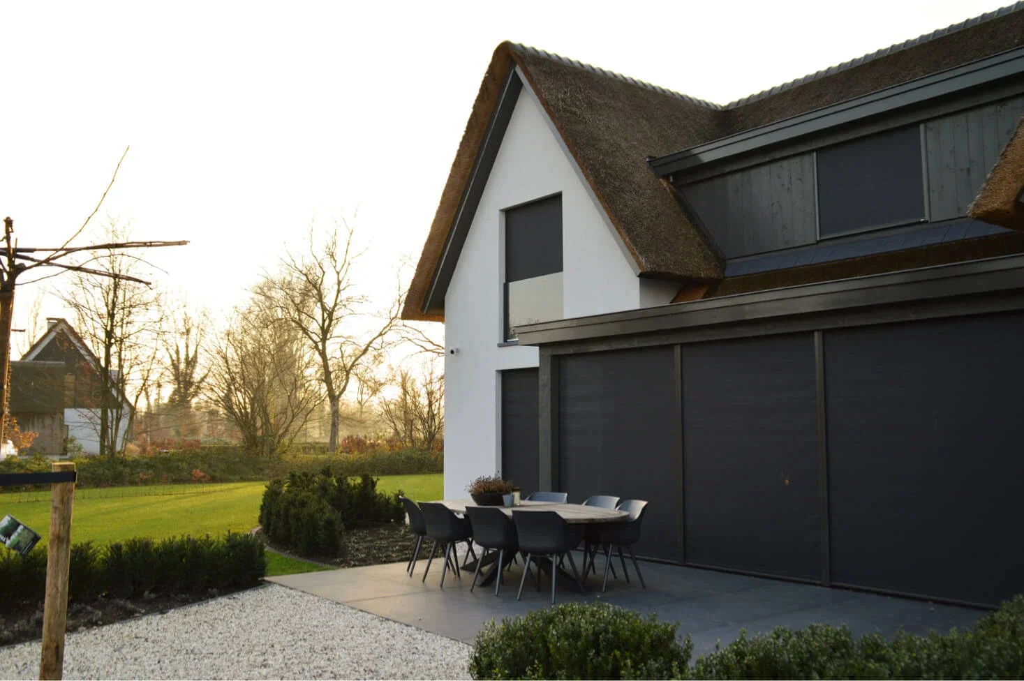 Moderne woning met een rieten dak en een terras met een zwart tafel en stoelen, omgeven door een tuin met groen gras en kleine planten, tijdens zonsondergang.
