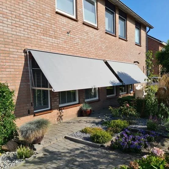 Witte uitvalschaduwdoek boven een tuin met planten en een stenen pad, bevestigd aan een bakstenen huisgebouw.