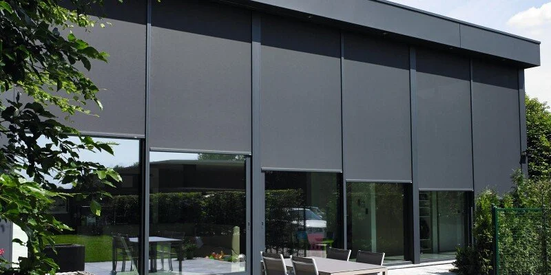 Modern zwart gebouw met grote glazen ramen en een patio met tafel en stoelen, omringd door groen tuin.