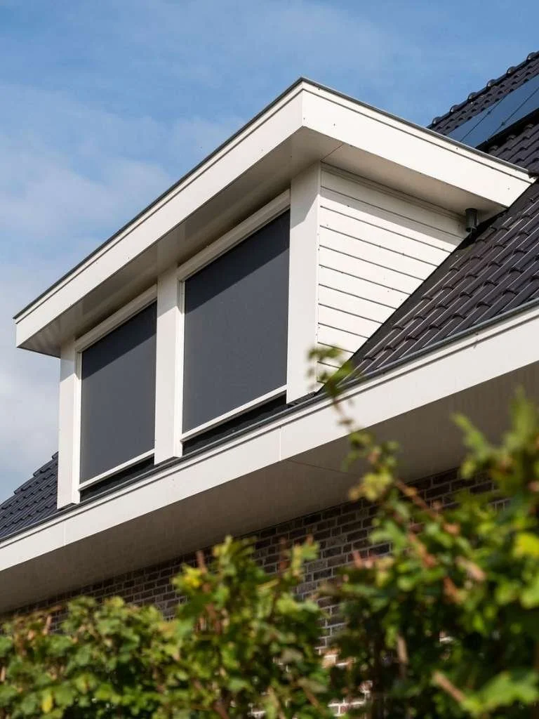 Uitzicht op een modern wit huis met drie grote ramen met zwarte rolgordijnen, een afgeplatte dakrand en een spantenstructuur, met groene planten onderaan en een blauwe hemel op de achtergrond.