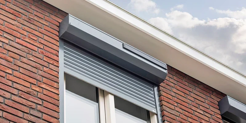 Natuurfoto van een raam met een metalen luifel en een houten bak boven een rode bakstenen muur, onder een bewolkte hemel.