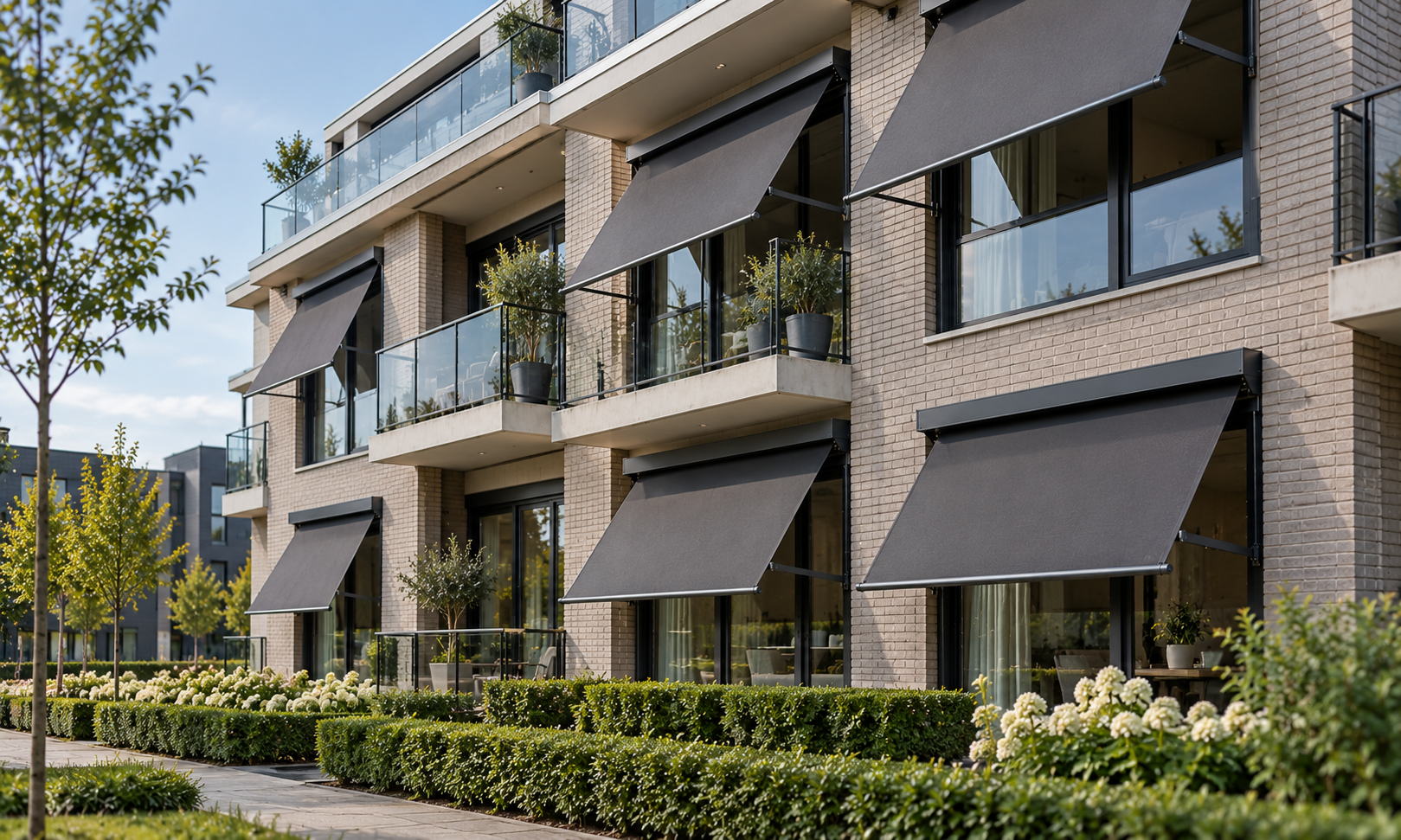A modern appartementgebouw met meerdere verdiepingen, grote ramen en zwarte uitvalschermen op de balkons, omgeven door groen gras en struiken.