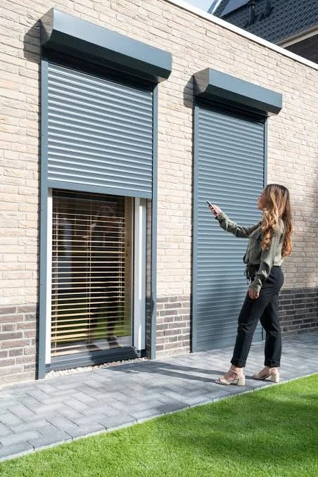 Vrouw bedient automatische rolluiken voor ramen aan een buitenmuur van een huis.