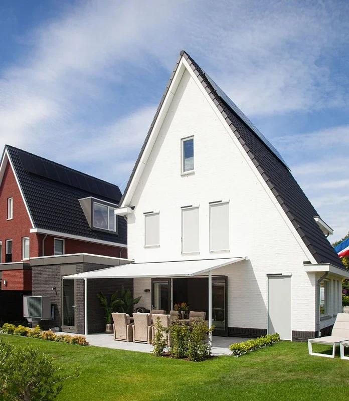 Moderne witte huis met een puntdak, terras met tuinmeubilair en een tuin, onder een blauwe lucht.