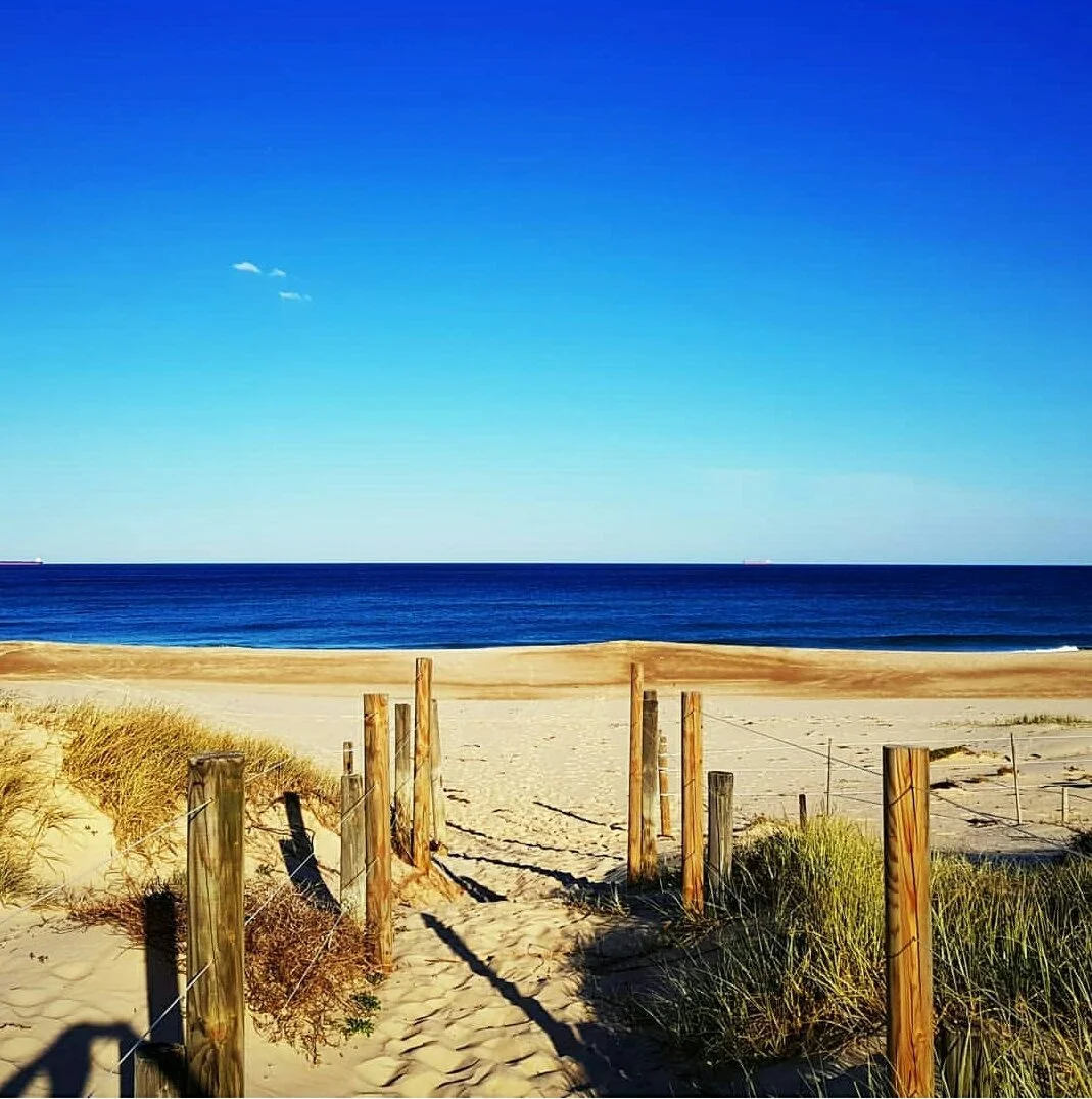 A sandy beach with a wooden pathway leading to the ocean, green beach grass on sides, clear blue sky with a few clouds.