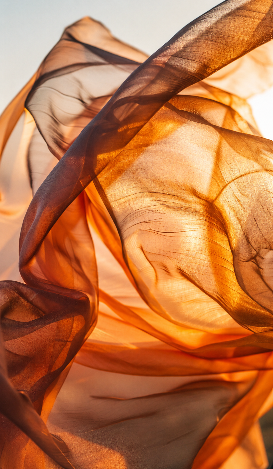 Close-up of flowing, translucent fabric in shades of orange, brown, and black with backlighting, creating abstract, wave-like patterns.