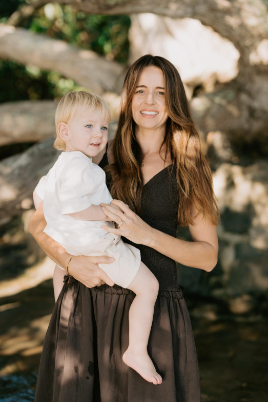 A woman with long brown hair holding a young girl with blonde hair outdoors near rocks and trees.