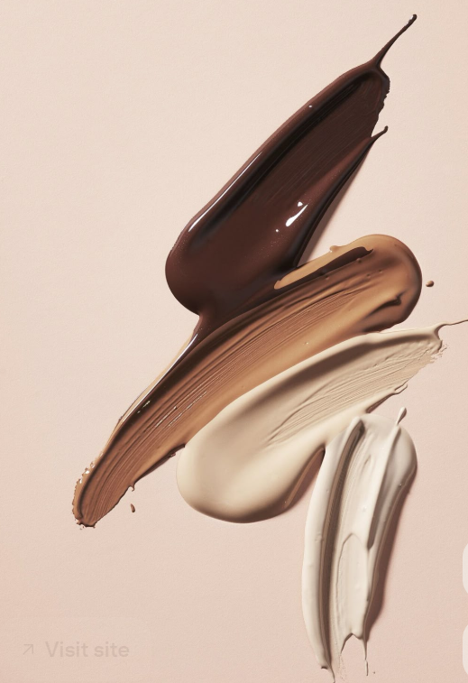 Swatches of different shades of foundation or concealer, including dark brown, light brown, beige, and white, spread on a light background.
