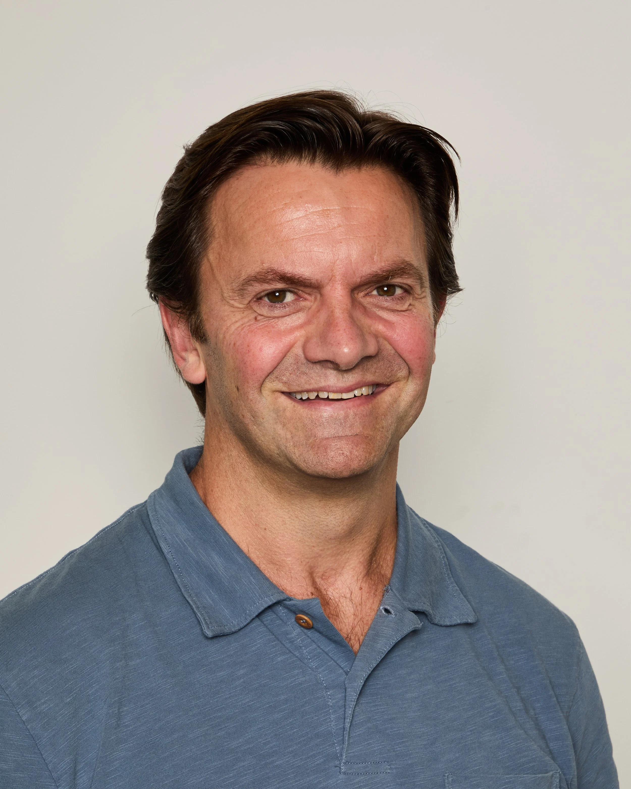Close-up of a smiling man with dark brown hair, wearing a blue collared shirt, against a plain light background.