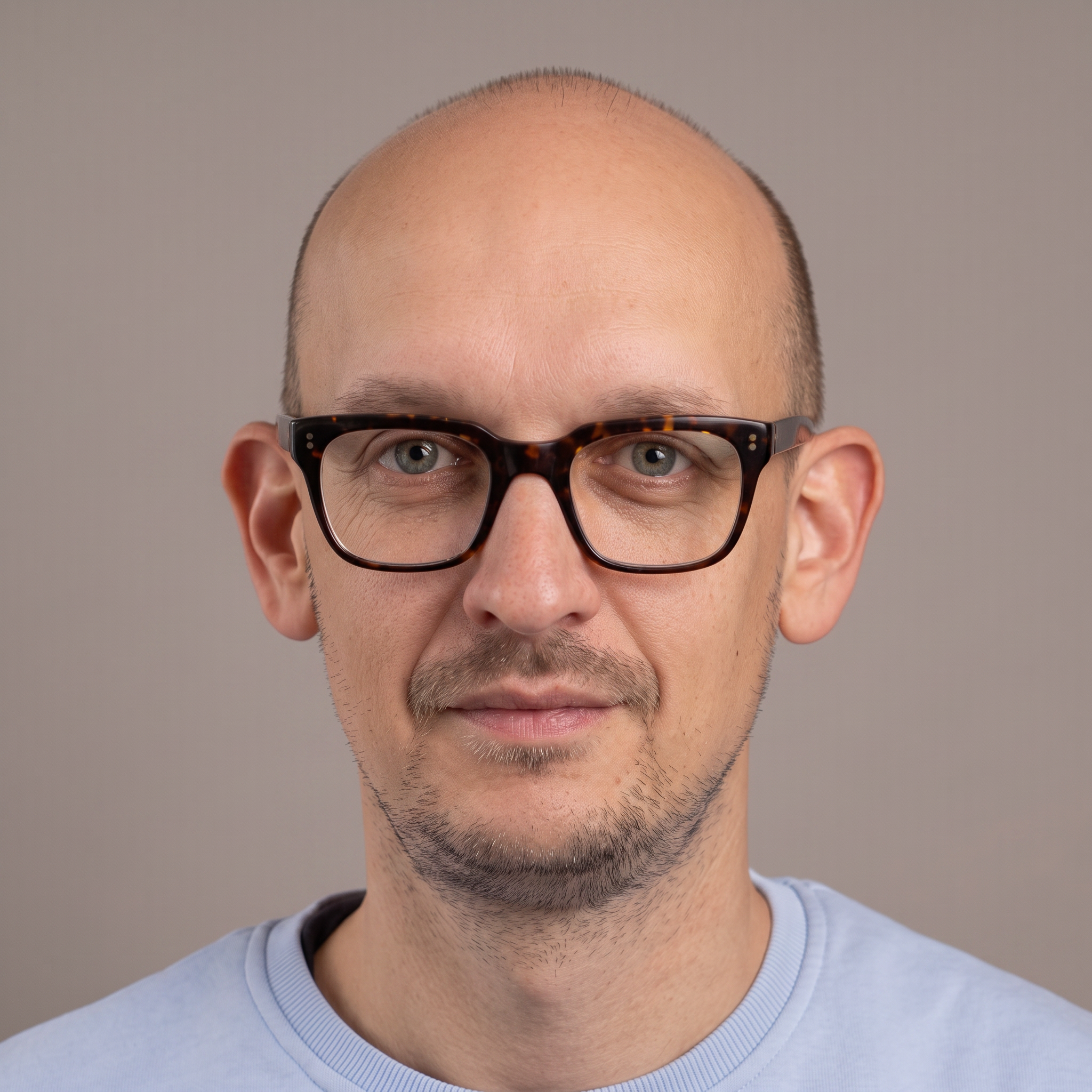 A close-up of a man's face showing a bald scalp, wearing tortoiseshell glasses, with light skin and a slight beard, against a plain gray background.