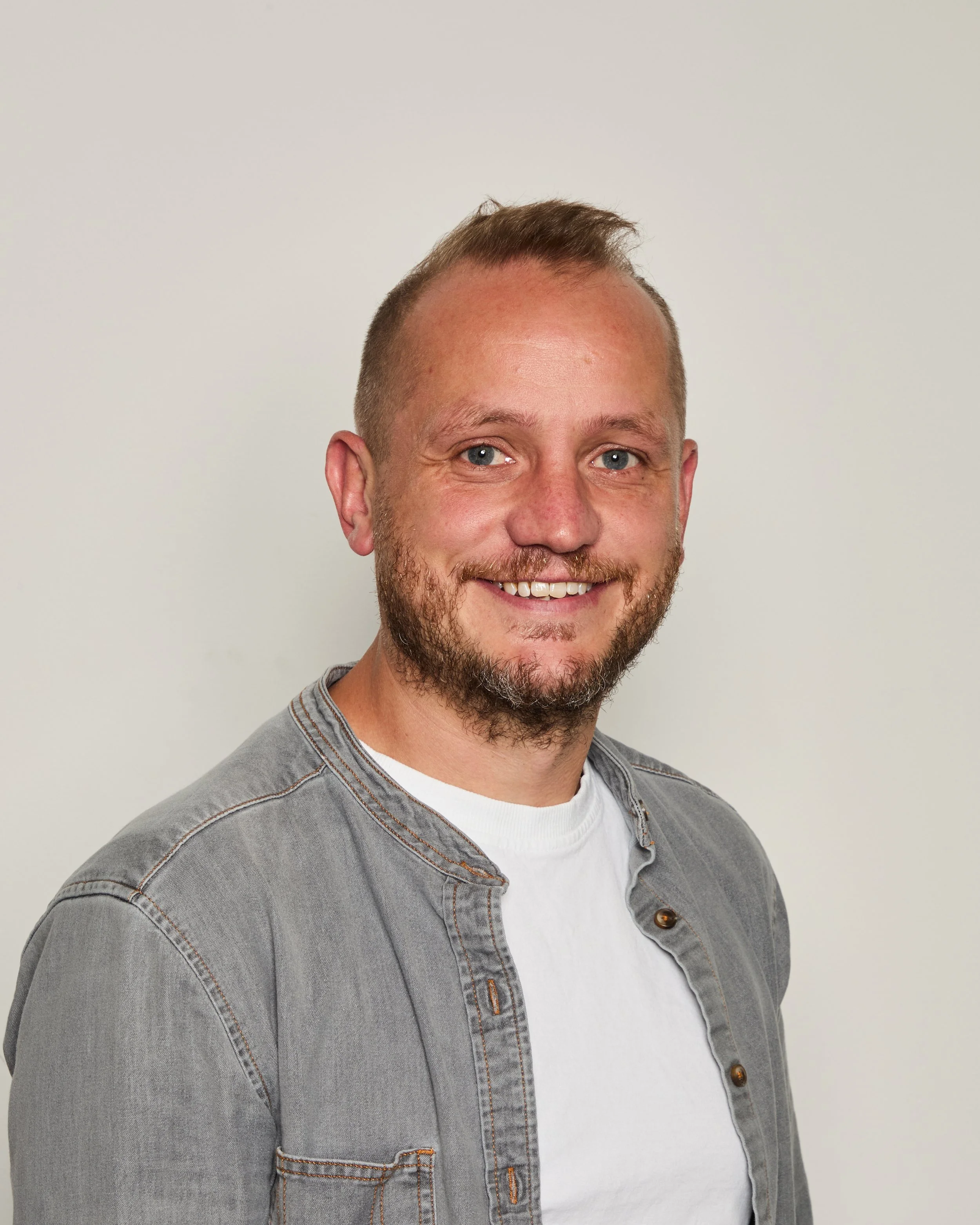 Close-up of a smiling man with short hair and a beard, wearing a grey denim jacket and a white t-shirt, standing against a plain white background.