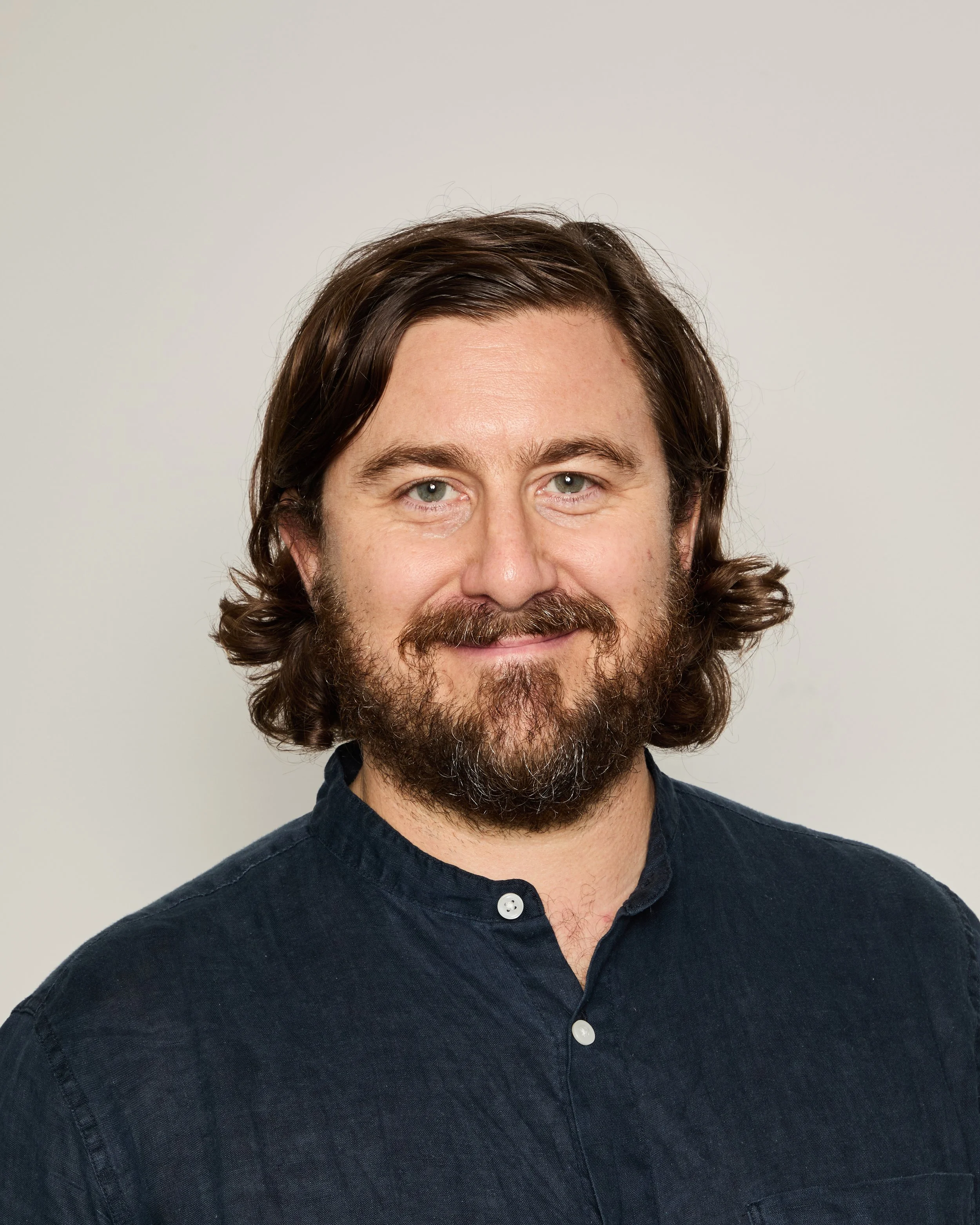 Close-up of a man with brown hair and a beard, wearing a dark button-up shirt, smiling gently against a plain light background.