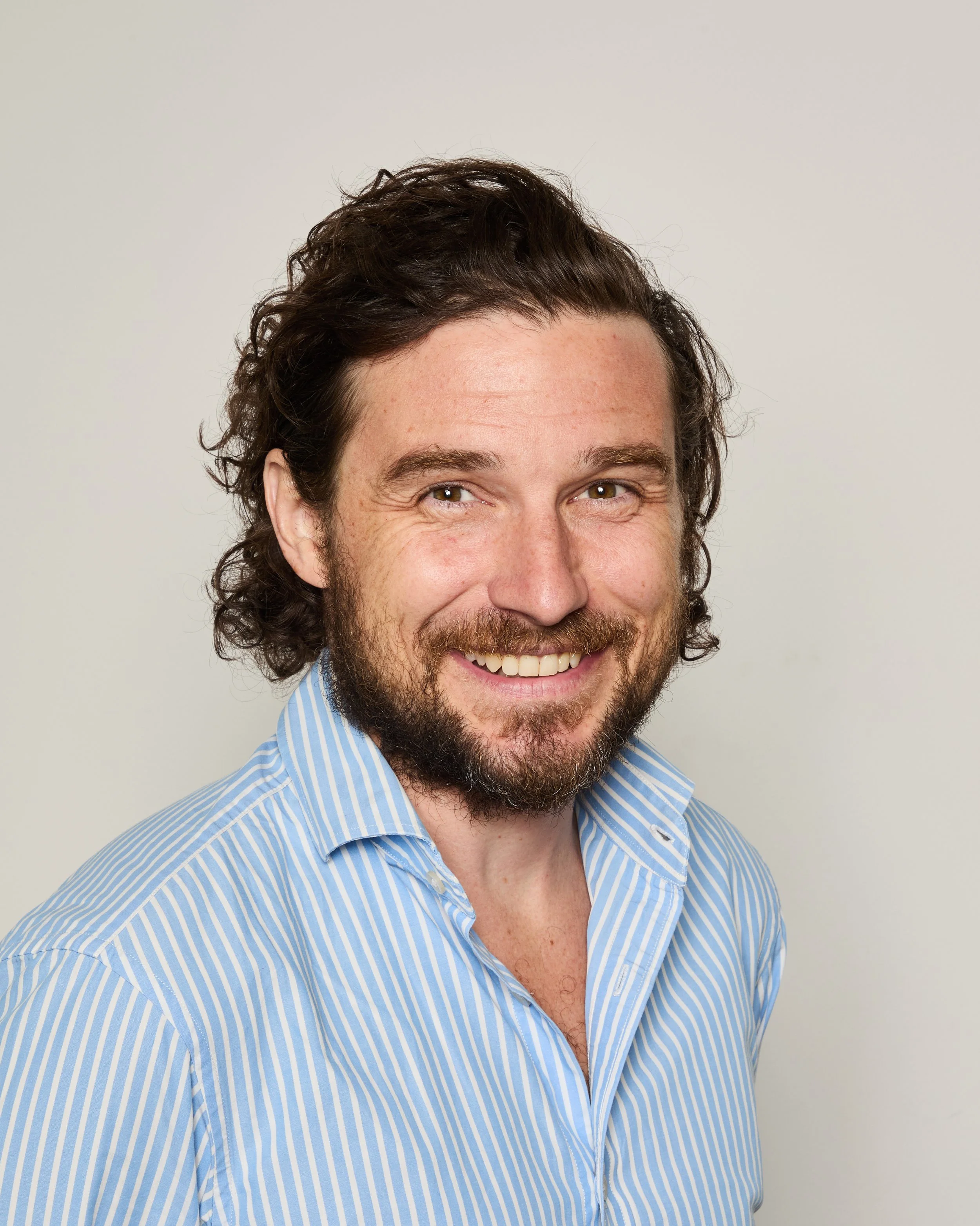 A smiling man with curly brown hair, a beard, wearing a blue and white striped shirt, against a plain white background.