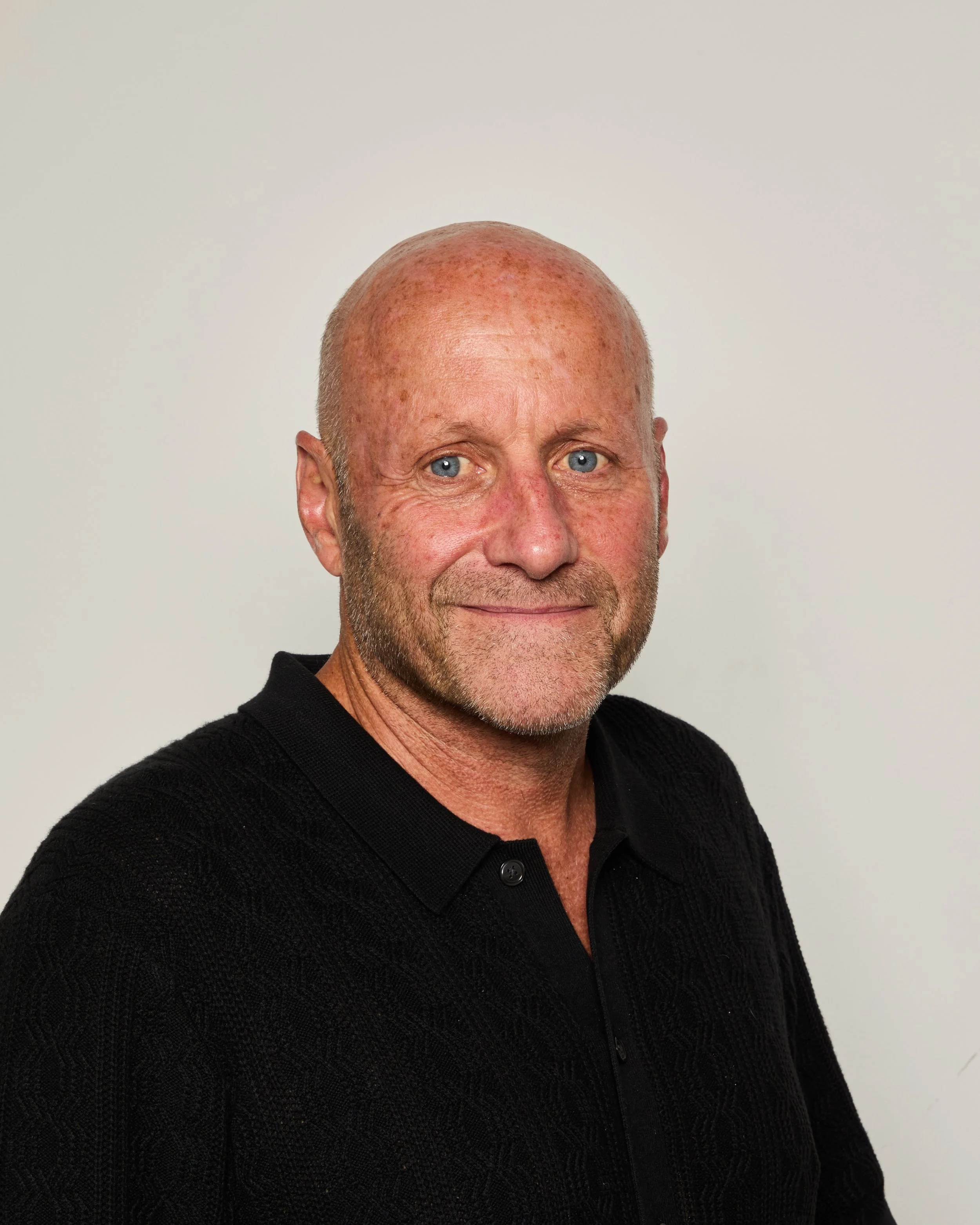 A middle-aged man with blue eyes and a bald head, wearing a black sweater, smiling softly against a plain light background.