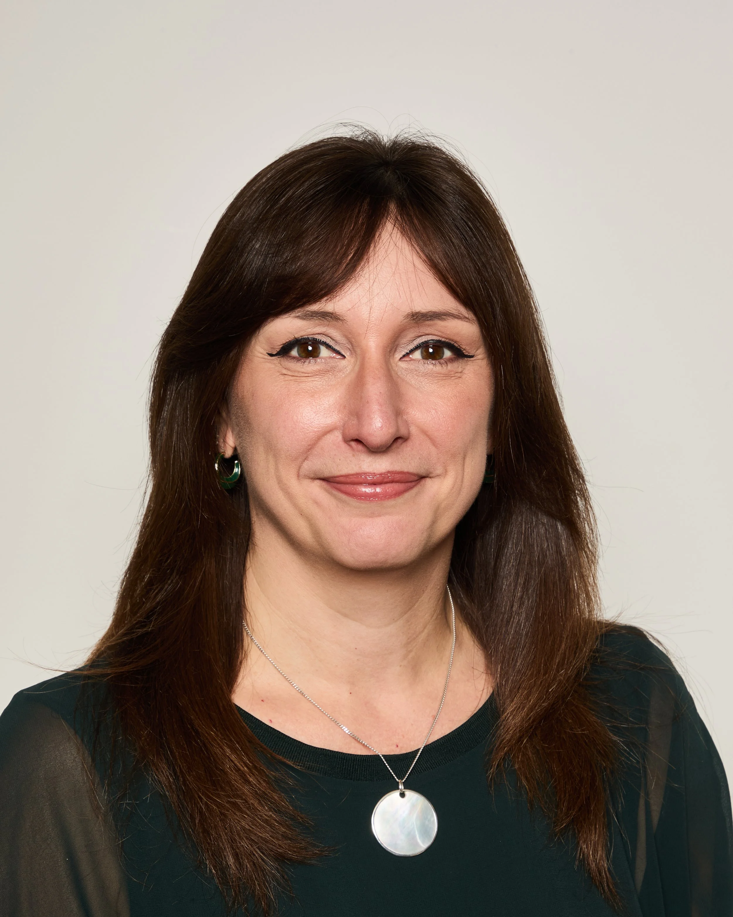 Close-up photo of a woman with brown hair, smiling slightly, wearing earrings, a necklace, and a black top.