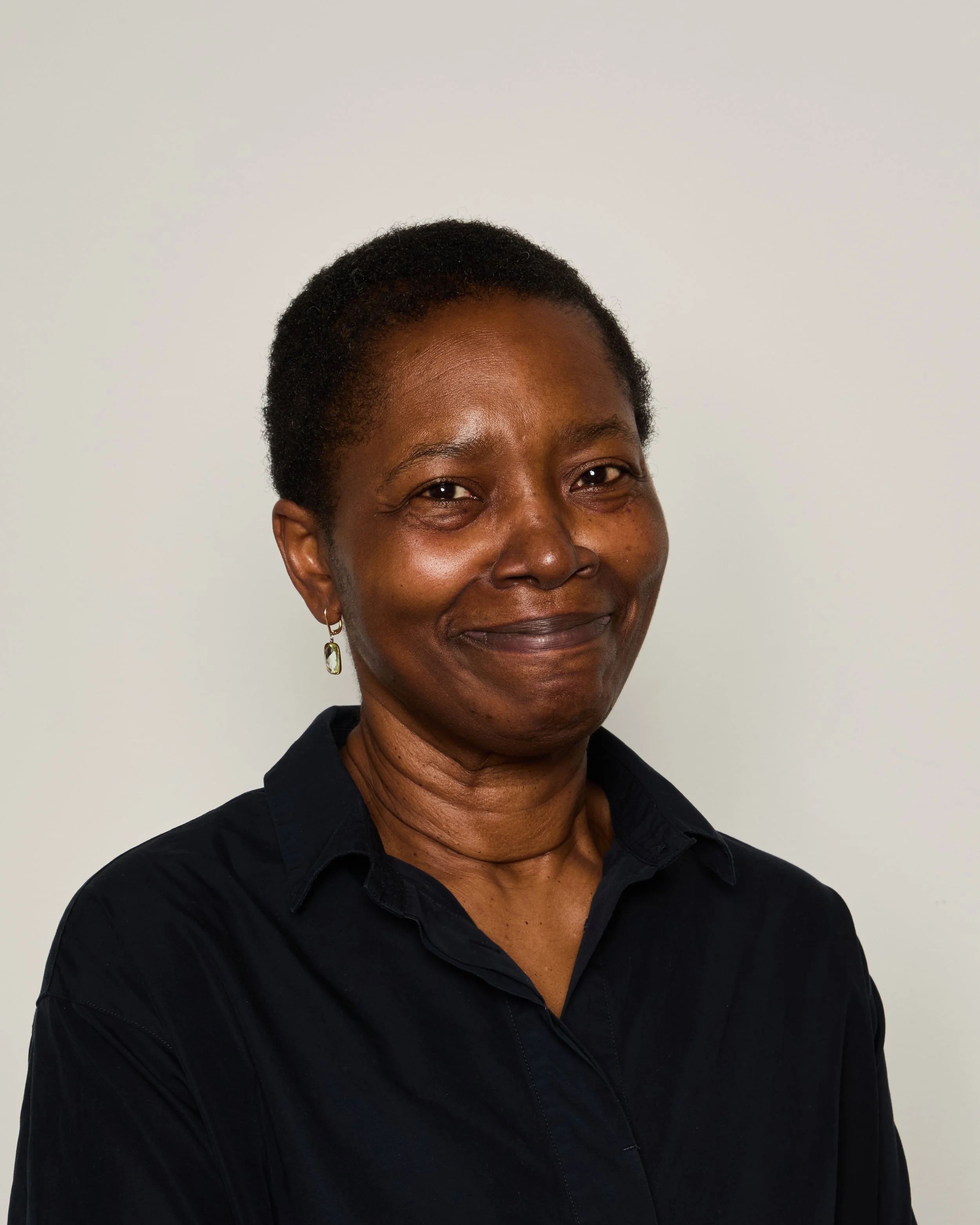 Close-up of a woman with short black hair, wearing earrings, smiling, against a plain white background, dressed in a black shirt.
