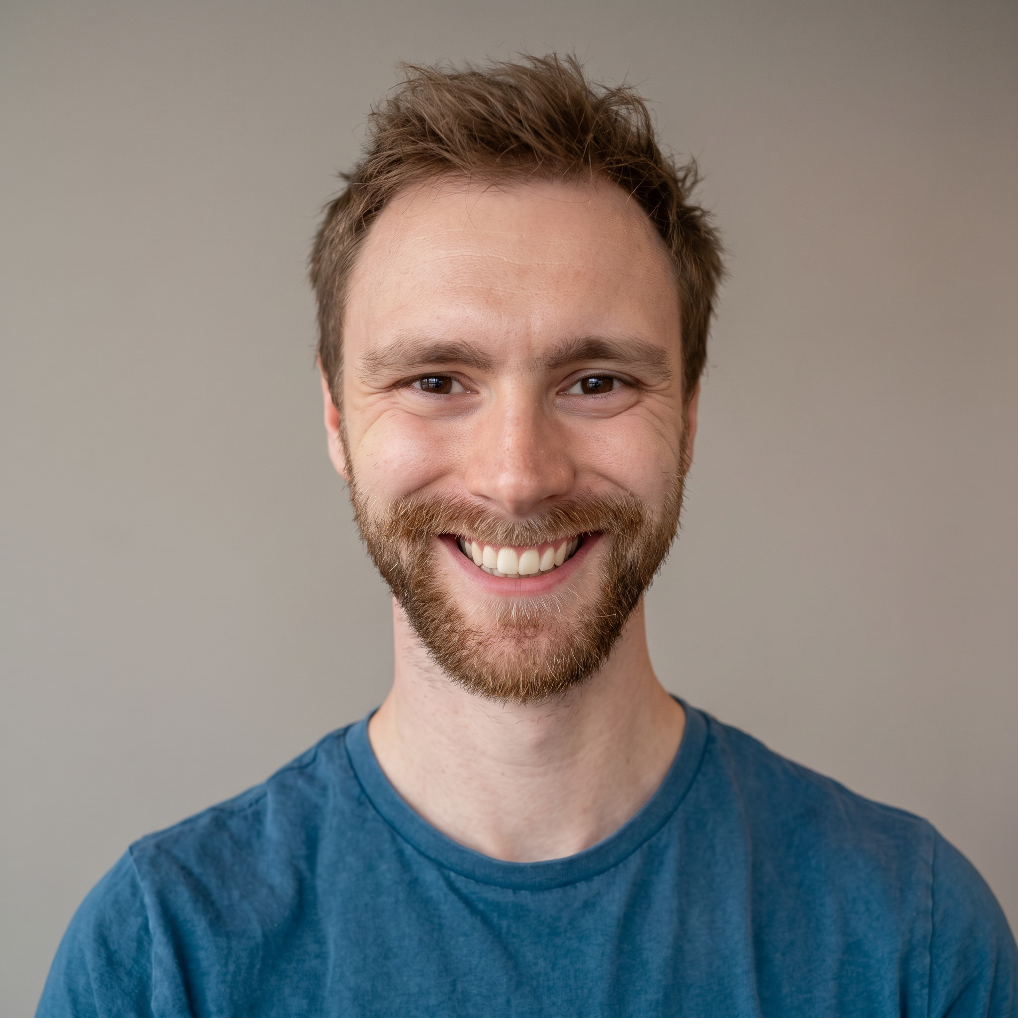 Close-up of a smiling young man with facial hair, wearing a blue shirt, against a neutral background.